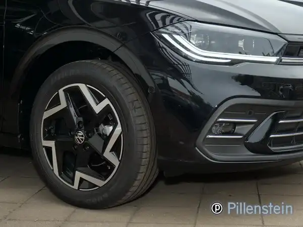 Das Bild zeigt die Vorderansicht eines schwarzen VW Polo. Das Fahrzeug ist teilweise sichtbar, insbesondere der Reifen und ein Teil der Karosserie, mit einem Händlerlogo im Bild.