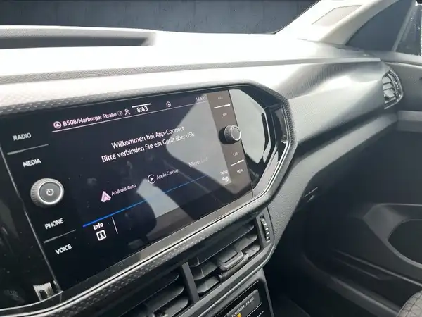 Das Bild zeigt das Innere eines VW T-Cross mit einem digitalen Bildschirm, der verschiedene Funktionen anzeigt. Das Interieur ist in schwarz gehalten.