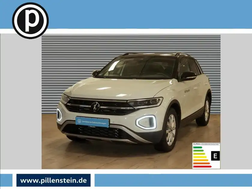 Das Bild zeigt einen weißen VW T-Roc von vorne, aufgenommen in einem Ausstellungsraum. Im Bild ist das Händlerlogo zu sehen und es gibt markante Texteinblendungen.