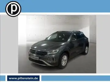 Das Bild zeigt einen VW T-ROC in grauer Farbe, aufgenommen von der Seite. Es sind das Händlerlogo und markante Texteinblendungen im Bild enthalten.