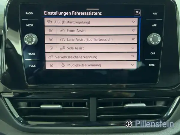 Eine Innenaufnahme des Fahrzeugs zeigt das Menü für Fahrsysteme auf einem Touchscreen, die Bedienelemente sind schwarz und das Bild enthält das Händlerlogo.