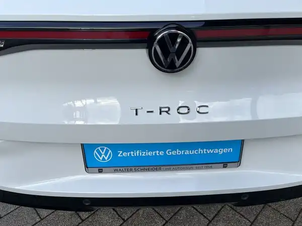 Das Bild zeigt die Rückansicht eines weißen VW T-Roc, mit einem Händlerlogo und einer markanten Texteinblendung für zertifizierte Gebrauchtwagen.