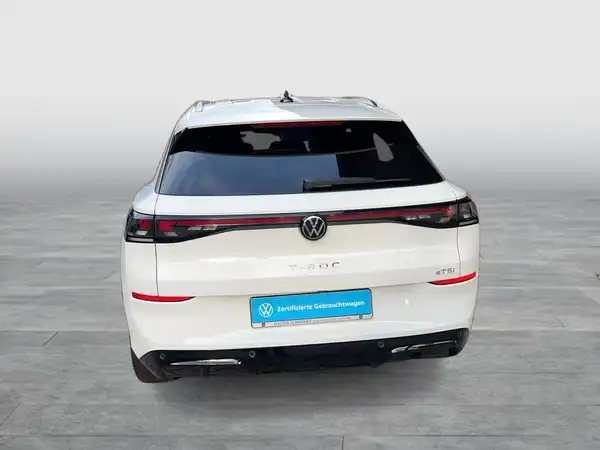 Das Bild zeigt die Rückansicht eines weißen Volkswagen T-Roc. Das Händlerlogo ist sichtbar und es gibt markante Texteinblendungen.