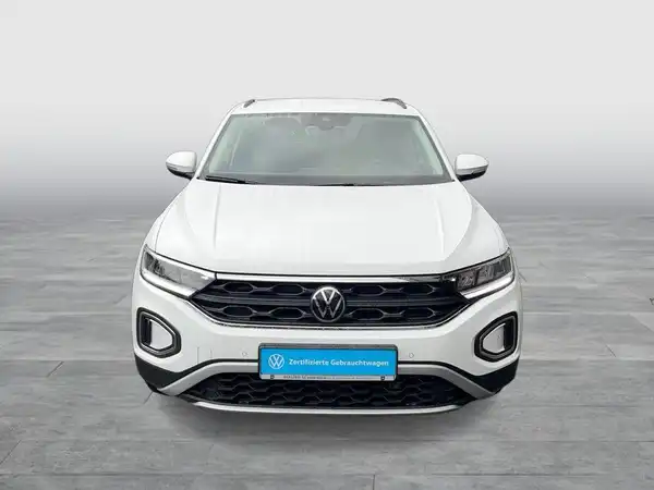 Das Bild zeigt einen weißen VW T-Roc von vorne. Im Bild ist das Händlerlogo sowie markante Texteinblendungen sichtbar.