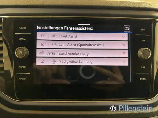 Das Bild zeigt das Innere eines VW T-ROC mit einem Display, auf dem die Einstellungen für den Fahrerassistenzsystem angezeigt werden. Die Innenfarbe scheint schwarz zu sein, und das Händlerlogo ist sichtbar.