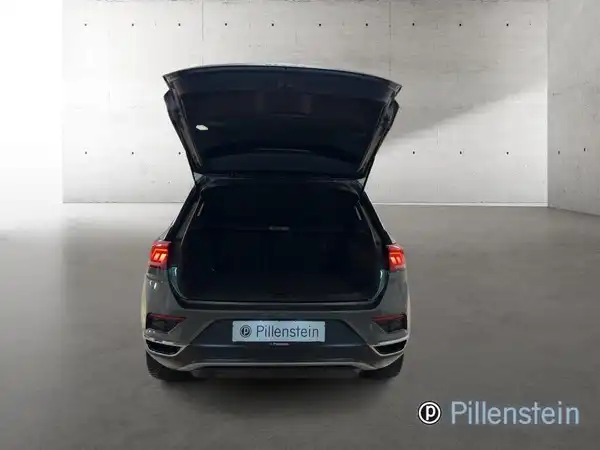 Das Bild zeigt die Rückansicht eines grauen VW T-ROC mit geöffnetem Kofferraum in einer neutralen Umgebung.