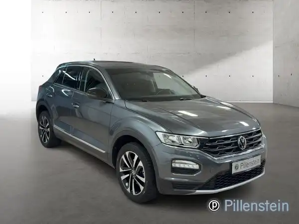 Das Bild zeigt einen grauen VW T-ROC von der Seite. Im Hintergrund sind minimale Texturen sichtbar, und das Händlerlogo ist auf dem Bild eingeblendet.