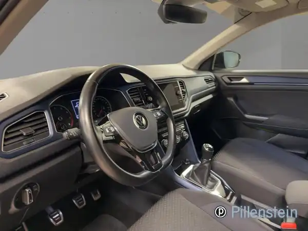 Das Bild zeigt das Innere eines Volkswagen T-ROC in schwarzem Interieur. Es enthält das Logo des Händlers.