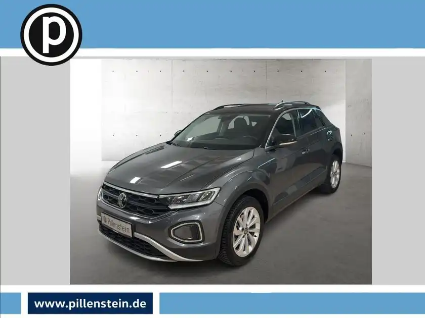 Das Bild zeigt einen grauen VW T-ROC, aufgenommen von der Seite. Es enthält das Händlerlogo und es sind markante Texteinblendungen zu sehen.