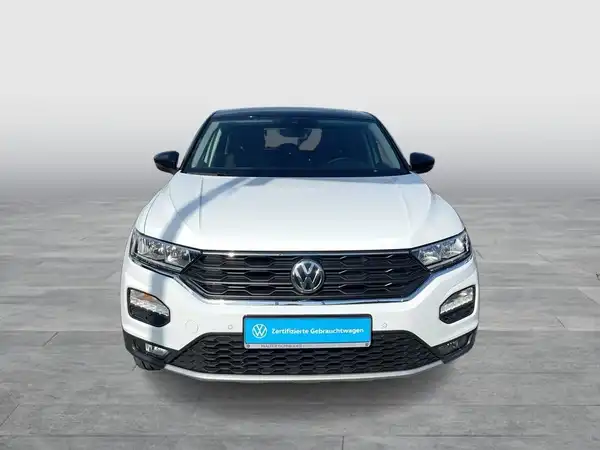 Das Bild zeigt einen weißen Volkswagen T-Roc von vorne. Es enthält ein Händlerlogo und eine markante Texteinblendung.