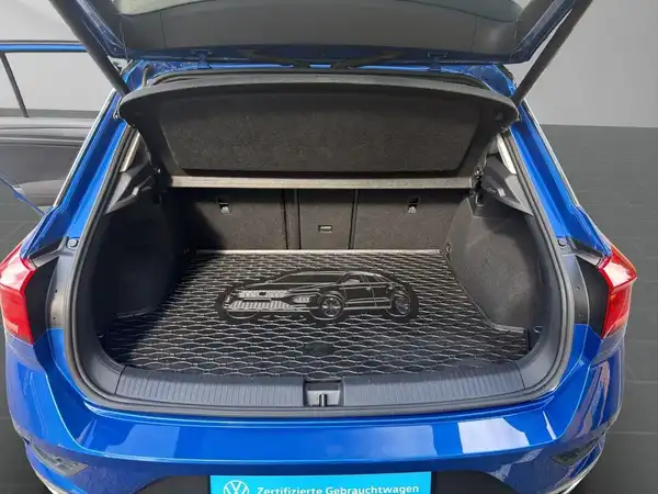 Das Bild zeigt den Kofferraum eines blaue VW T-Roc von hinten. Der Kofferraumdeckel ist geöffnet und im Kofferraum ist eine Abdeckung zu sehen.