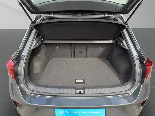 Das Bild zeigt den Kofferraum eines grauen Volkswagen T-Roc aus der Rückansicht. Es enthält ein Händlermarkenzeichen.