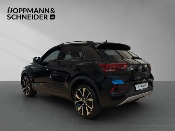 Das Bild zeigt einen schwarzen VW T-Roc von hinten. Im Hintergrund ist das Händlerlogo "HOPPMANN & SCHNEIDER" zu sehen.