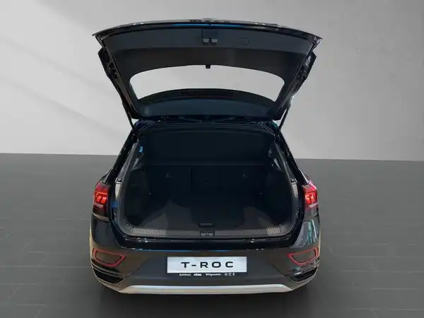 Das Bild zeigt einen VW T-Roc von hinten mit geöffnetem Kofferraum. Das Fahrzeug ist schwarz.