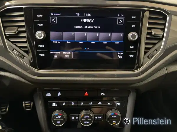 Das Bild zeigt das Innenraum des VW T-ROC, dabei ist das schwarze Interieur und das Bedienfeld mit einem Display sichtbar. Es ist das Händlerlogo von Pillenstein eingeblendet.