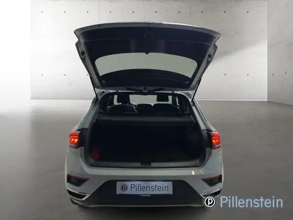 Das Bild zeigt die Rückansicht eines weißen VW T-Roc mit geöffnetem Kofferraum. Im Hintergrund sind helle Wände zu sehen. Das Händlerlogo 'Pillenstein' ist eingeblendet.