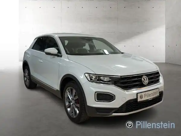 Das Bild zeigt einen VW T-Roc in der Farbe weiß, aufgenommen von der Seite. Es enthält das Händlerlogo von Pillenstein.