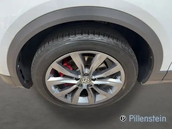 Das Bild zeigt einen VW T-ROC von der Seite, mit einem weißen Karosseriefinish und einem Blick auf das Rad und die Felge. Auf dem Foto ist das Händlerlogo von Pillenstein eingeblendet.