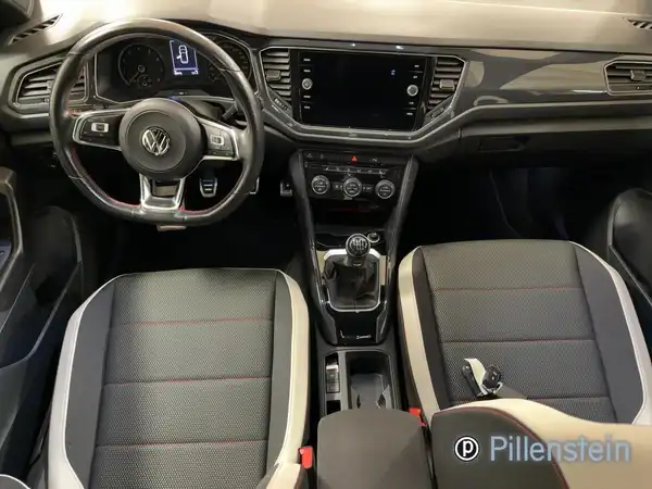Das Bild zeigt das Innere eines VW T-ROC mit schwarzen Sitzen und grauen Akzenten. Es gibt ein Händlerinlogo auf dem Bild.
