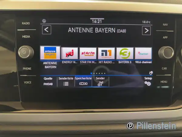 Das Bild zeigt das Innere eines VW Taigo mit einem Navigations- und Multimedia-Bildschirm, der verschiedene Radiostationen anzeigt. Der Bildschirm hat ein schwarzes Interieur.