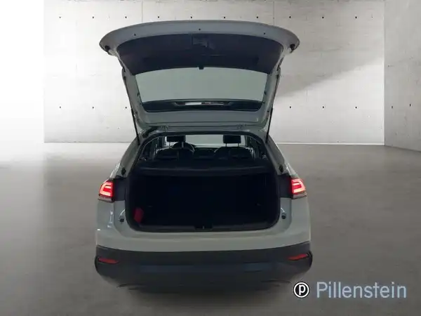 Das Bild zeigt die Heckansicht eines weißen VW Taigo mit geöffnetem Kofferraum. Im Hintergrund ist eine neutrale Wand zu sehen. Ein Händlermarkenzeichen ist sichtbar, jedoch keine markanten Texteinblendungen.