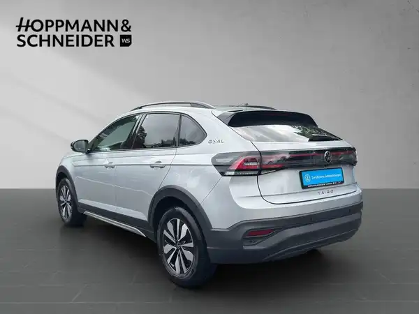 Das Bild zeigt einen VW Taigo von der Seite. Das Fahrzeug ist silberfarben. Darüber hinaus ist das Händlerlogo "HOPPMANN & SCHNEIDER" sichtbar.