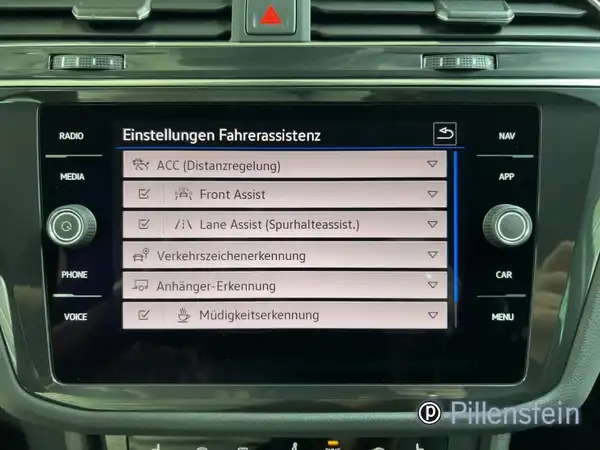 Das Bild zeigt den Innenraum eines VW Tiguan, mit Fokus auf das Display des Infotainment-Systems, das Einstellungen für den Fahrassistenten anzeigt. Das Interieur ist schwarz und es befindet sich ein Händlerlogo auf dem Bild.