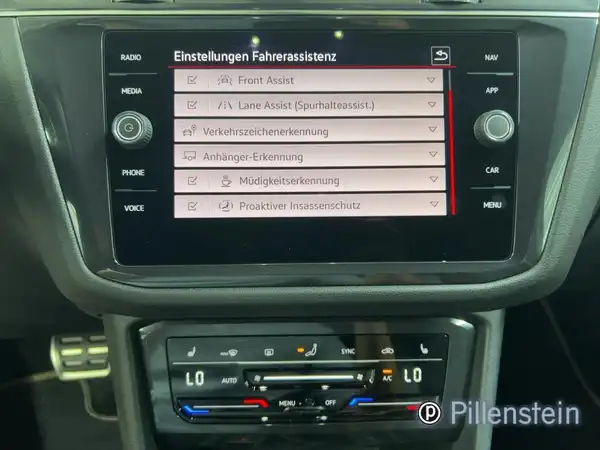 Das Bild zeigt das Innere eines VW Tiguan Allspace mit einem Dashboard, von dem die Einstellungen für die Fahrassistenz angezeigt werden. Die Innenfarbe ist schwarz, und es gibt ein Händlerlogo auf dem Bild.