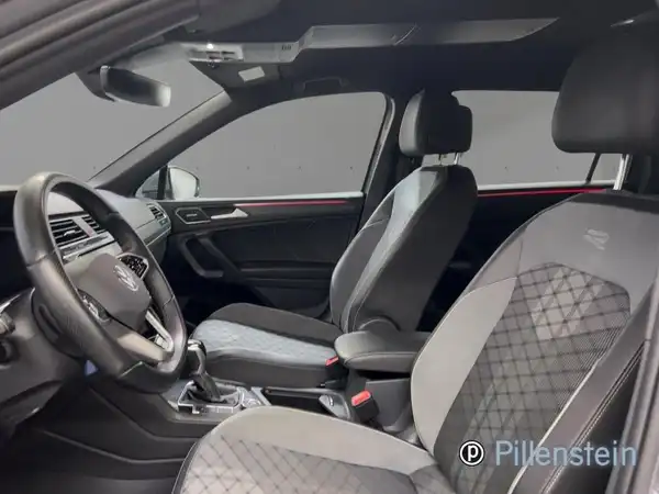 Foto zeigt das Innenraum eines VW TIGUAN ALLSPACE mit sportlichen schwarzen Sitzen und geometrischem Muster. Händlerlogo ist sichtbar.