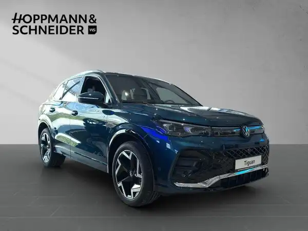 Dies ist ein Bild eines blauen VW Tiguan, aufgenommen von der Seite. Das Fahrzeug ist in einem modernen Ausstellungsraum platziert, mit dem Händlerlogo 'HOPPMANN & SCHNEIDER' im Hintergrund.