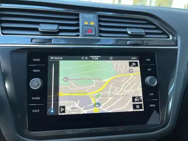 Das Bild zeigt das Innere eines VW Tiguan Allspace, insbesondere das Navigationssystem mit einer Karte, die Informationen zur Umgebung anzeigt. Das Interieur ist schwarz.
