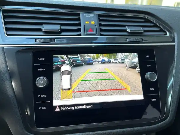 Das Bild zeigt das Innenraumdisplay eines VW Tiguan Allspace mit einer Rückfahrkamera-Anzeige. Der Bildschirm ist schwarz und zeigt die Umgebung hinter dem Fahrzeug an, mit Hilfslinien in Grün, Gelb und Rot. Es ist eine Innenaufnahme.