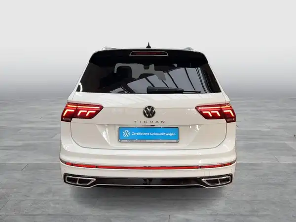 Das Bild zeigt einen weißen VW Tiguan Allspace von hinten, mit einem sichtbaren Händlerlogo und markanten Texteinblendungen.