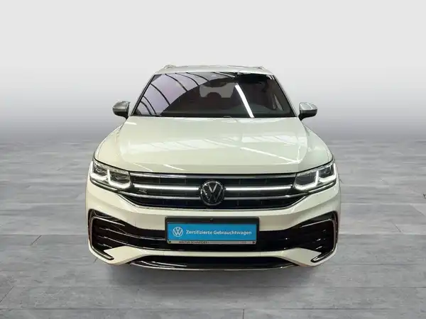 Das Bild zeigt einen weißen VW Tiguan Allspace von vorne. Auf dem Bild ist das Händlerlogo eingeblendet und es enthält markante Texteinblendungen.