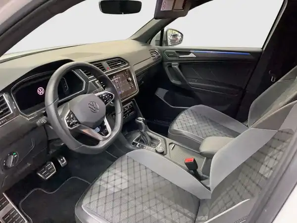 Das Bild zeigt das Innere eines VW Tiguan Allspace, aufgenommen aus einem Blickwinkel, der das Lenkrad, das Armaturenbrett und die Vordersitze zeigt. Die Innenausstattung ist grau mit einem Karomuster.