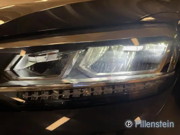 Das Bild zeigt die Frontansicht des Scheinwerfers eines schwarzen VW Touran. Es ist ein Bild des Fahrzeugs, auf dem das Händlerlogo zu sehen ist.
