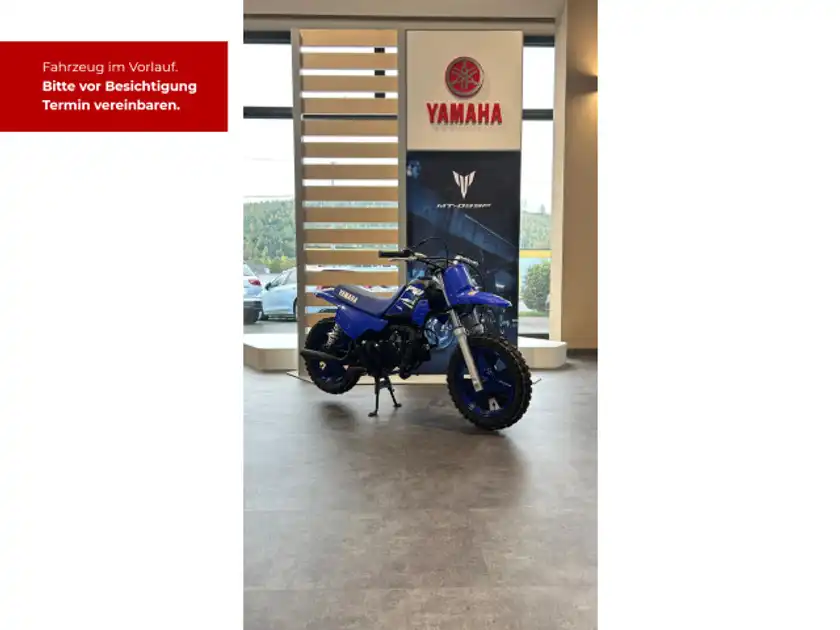 Das Bild zeigt ein YAMAHA PW50 aus der Seitenansicht in blauer Farbe. Im Hintergrund sind Werbeanzeigen zu sehen mit einem Händlerinformationstext und dem Händlerlogo.