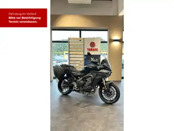 Das Bild zeigt eine schwarze Yamaha Tracer 9 aus einer Seitenansicht. Im Hintergrund sind Hinweise für eine Besichtigung sowie ein Händlerschild zu sehen.
