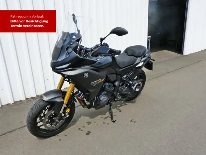 Das Bild zeigt eine schwarze Yamaha Tracer von der Seite. Im Hintergrund ist eine weiße Wand zu sehen, und es gibt markante Texteinblendungen in rot, die auf eine Fahrzeugbesichtigung hinweisen.