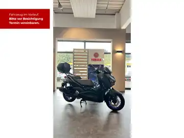 Das Bild zeigt ein schwarzes YAMAHA XMAX 300 Motorfahrzeug, aufgenommen von der Seite in einem Ausstellungsraum. Im Bild sind Texteinblendungen zu sehen, die zur Terminvereinbarung auffordern, sowie ein Händlerlogo im Hintergrund.