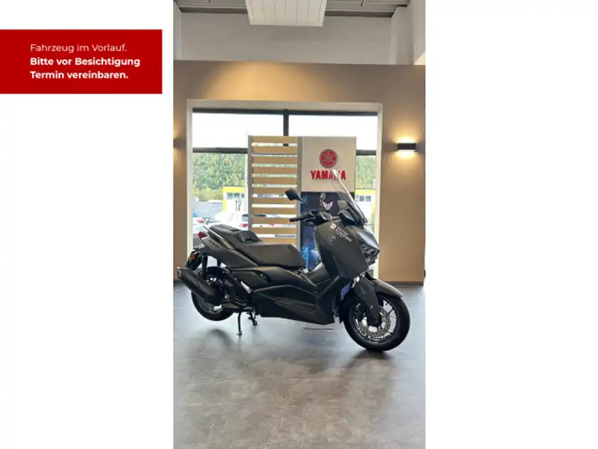 Bild zeigt einen schwarzen Yamaha XMAX 300 aus seitlicher Perspektive in einem Verkaufsraum, mit Händlerlogo und markantem Text, der zur Terminvereinbarung auffordert.