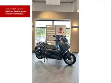 Bild zeigt einen schwarzen Yamaha XMAX 300 aus seitlicher Perspektive in einem Verkaufsraum, mit Händlerlogo und markantem Text, der zur Terminvereinbarung auffordert.
